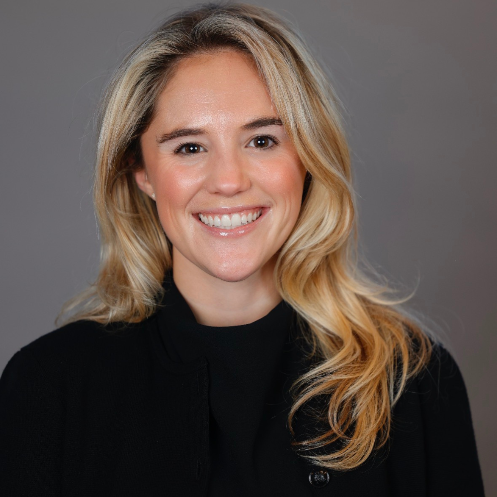 Professional headshot of a smiling woman with blonde hair wearing a black top against a gray background